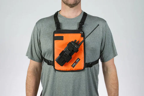 A person wears a gray t-shirt and a Lone Peak Packs Universal Radio Chest Pack in orange, holding a black two-way radio. The harness secures with black straps around the shoulders and chest.