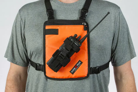 A person in a gray t-shirt wears a Lone Peak Packs Universal Radio Chest Pack in orange, strapped around the shoulders and torso, with a black radio secured in the front pocket.