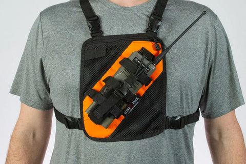 A person wearing a gray shirt and the Lone Peak Packs Universal Radio Chest Pack, featuring an orange and black radio holder, securely carries a handheld radio with antenna.