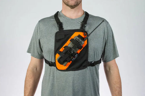 A person wears a gray t-shirt and carries a Lone Peak Packs Universal Radio Chest Pack in black and orange, secured with adjustable straps and designed to hold a handheld radio device on the chest.