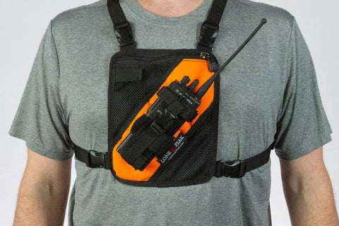 A person in a grey t-shirt wears the Lone Peak Packs Universal Radio Chest Pack, featuring black and orange design, adjustable radio pouch, and front straps to securely hold a walkie-talkie.