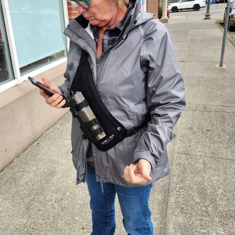A person in a gray jacket and blue jeans stands on the sidewalk, checking their phone. They carry a black Lone Peak Packs Universal Sling Radio Holster with a large camera lens attached. Parked cars and a building appear in the background.