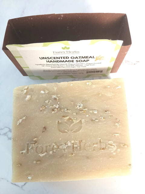 A bar of Forev'Herbs Unscented Oatmeal Handmade Soap, with visible oatmeal flakes and embossed logo, sits before its brown and white box—a moisturizing cleanser made to gently nourish and soothe dry skin.