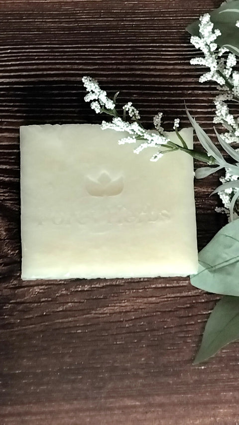 A square bar of Unscented Shea Handmade Soap by Forev'Herbs, with a subtle logo and text, rests on dark wood beside green leaves and white flowers—perfect for sensitive skin.