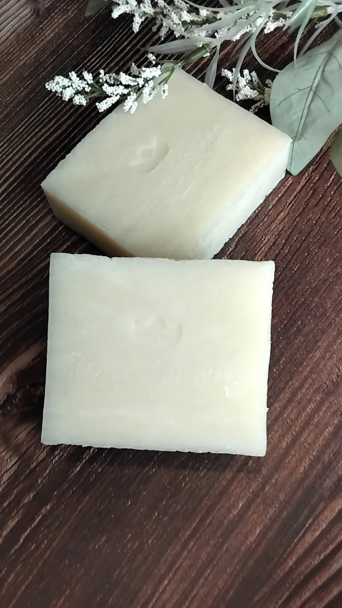 Two rectangular bars of Forev'Herbs Unscented Shea Handmade Soap with embossed text rest on a dark wooden surface, surrounded by dried flowers and green leaves—a natural, rustic setting ideal for sensitive skin care routines.