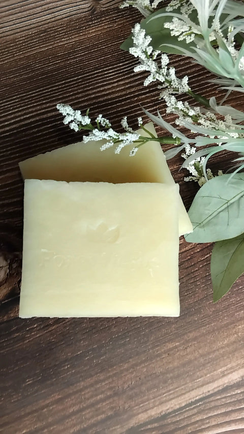 Two rectangular, pale yellow Forev'Herbs Unscented Shea Handmade Soap bars rest on wood with green leaves and small white flowers nearby—ideal for sensitive skin.