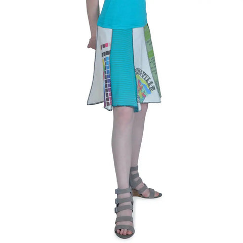 A person wears a turquoise top, gray strappy sandals, and the Upcycled Short Bell Skirt by Spiritex | Sustainable Apparel, featuring colorful patchwork designs, standing with one arm behind their back.