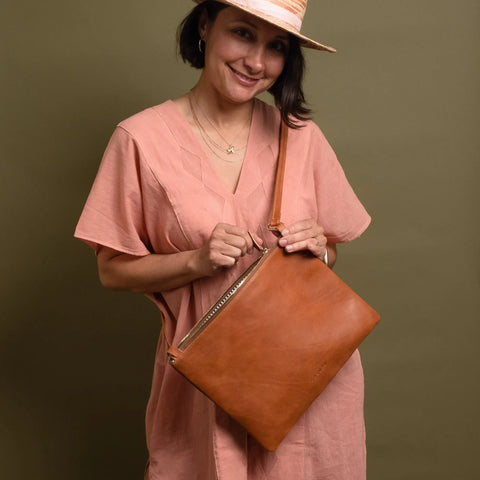 A woman in a light pink dress and hat smiles while holding the CASUPO Crossbody Bag - Natural, handcrafted from vegetable tanned leather, against an olive green background.