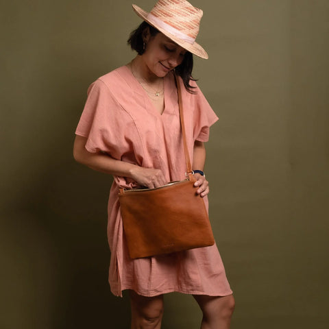 A woman in a light pink dress and straw hat looks down as she reaches into the CASUPO Crossbody Bag - Natural, made of handcrafted vegetable-tanned leather, set against an olive green background.