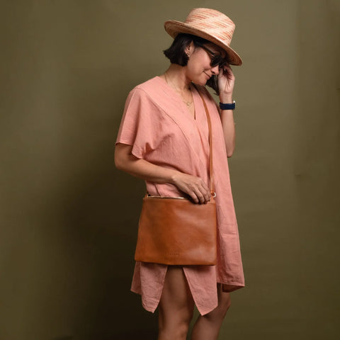 A woman in a pink dress, straw hat, and sunglasses stands against a plain background, holding the CASUPO Crossbody Bag - Natural and touching her face with one hand.