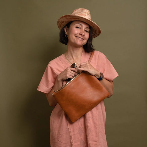 A woman in a pink dress and straw hat smiles while holding the CASUPO Crossbody Bag - Natural against a plain olive green background.