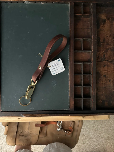The Sully + Proper Supply Key Keeper With Brass Alligator Clasp Hardware rests on a dark green pad atop a wooden desk. A white tag, part of a tan leather bag, and beige shorts are also visible in the scene.