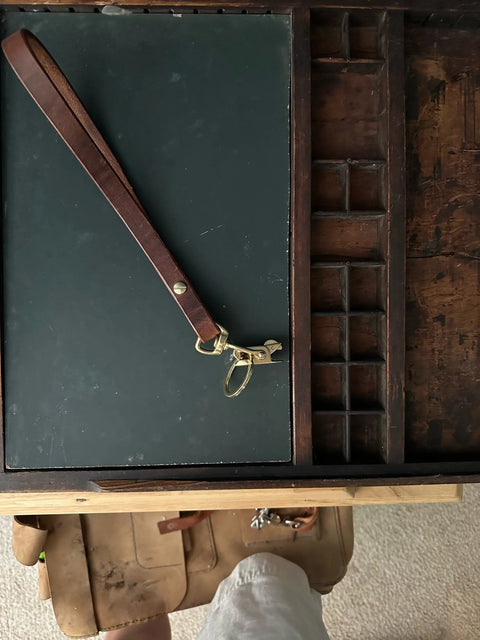 A Sully + Proper Supply Key Keeper with brass alligator clasp hardware rests on a dark surface in an open wooden drawer organizer. At the bottom, part of a person’s leg in shorts and shoes is visible.