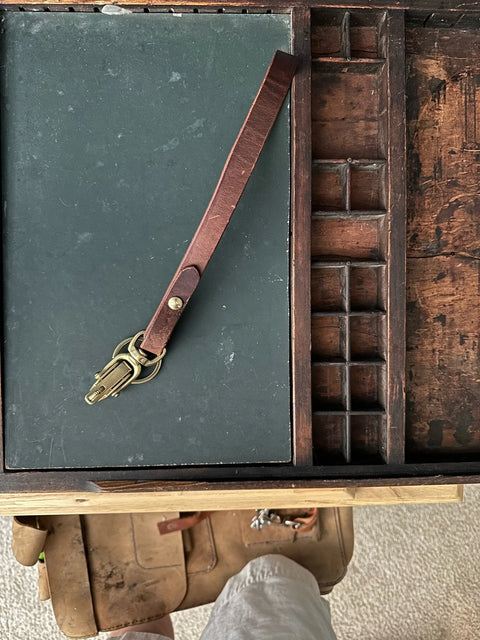 The Sully + Proper Supply Key Keepers With Brass Alligator Clasp Hardware rests on a dark green surface in an open, vintage wooden desk organizer, with part of a person’s legs and tool belt below—a nod to American-made craftsmanship.