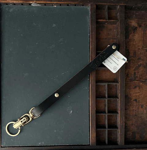 The Sully + Proper Supply Key Keepers With Brass Alligator Clasp Hardware in black vegetable-tanned leather sits diagonally on a closed dark green book inside a wooden tray, with a small white tag attached to the keychain.