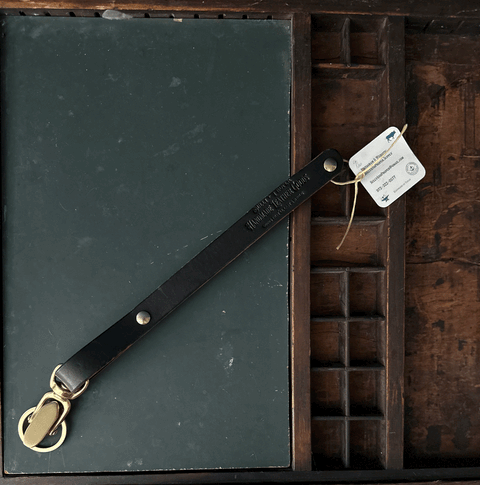 A Sully + Proper Supply Key Keeper with brass alligator clasp hardware rests diagonally across a dark green notebook inside a wooden compartment tray, showcasing American-made craftsmanship and featuring a white tag tied with string.