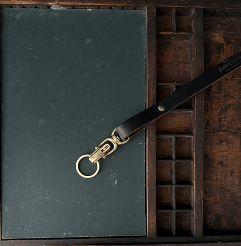 A closed dark green notebook and a Sully + Proper Supply Key Keeper with brass alligator clasp hardware rest in a wooden compartment tray, showcasing American-made craftsmanship.