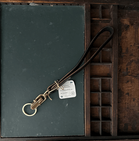 A green book with a Sully + Proper Supply Key Keeper, featuring brass alligator clasp hardware and a tag, rests on a wooden organizer with small compartments.