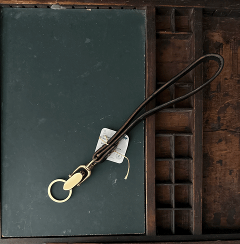 The Sully + Proper Supply Key Keeper features a vegetable-tanned leather strap, brass alligator clasp hardware, and a keyring. Made in the USA, it includes a small white tag and is shown on a dark green surface beside a wooden organizer.