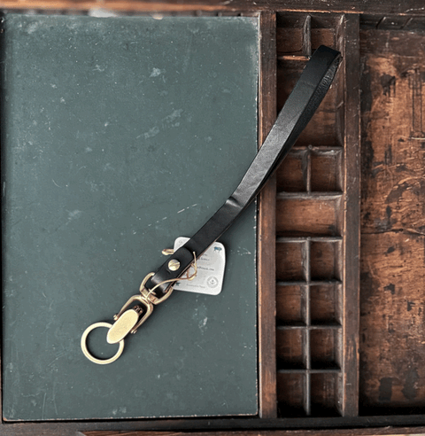 The Sully + Proper Supply Key Keepers With Brass Alligator Clasp Hardware rests on a closed dark green book, set inside a wooden compartmentalized organizer tray.