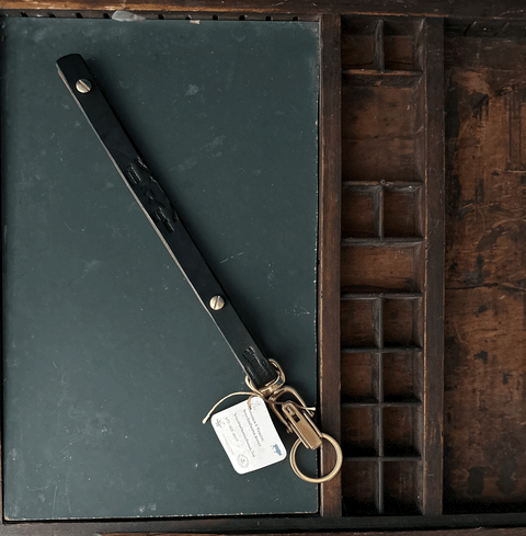 A Sully + Proper Supply Key Keeper with brass alligator clasp hardware and a white tag sits on a closed green book in a wooden drawer organizer with small compartments.
