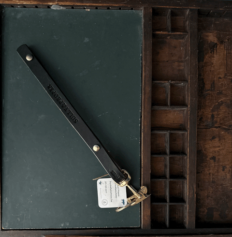 A Key Keeper With Brass Alligator Clasp Hardware by Sully + Proper Supply rests on a closed dark green notebook, all placed inside a wooden compartmentalized tray.