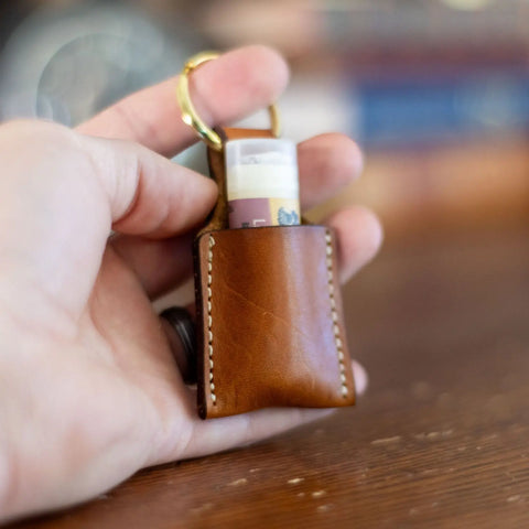 A hand holds the KC Laser Co Vegetable-Tanned Leather Lip Balm Holder, handmade in the USA, featuring visible stitching, a rolled banknote inside, and attached to a gold keyring against a blurred wooden background.