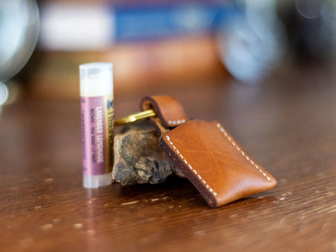 A tube of lavender-lemon lip balm, a small rock, and a KC Laser Co Vegetable-Tanned Leather Lip Balm Holder—handmade in the USA—rest on a wooden surface with blurred books in the background.
