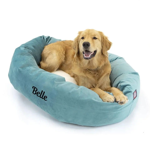 A golden retriever relaxes in a blue Velvet Sherpa Bagel Dog Bed by Majestic Pet, personalized with Belle embroidered on the side. The dog looks forward with its mouth open, appearing relaxed and comfortable.