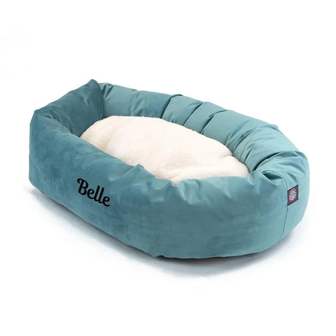 The Majestic Pet Velvet Sherpa Bagel Dog Bed - Personalized features a teal, oval shape with a soft white cushion. Belle is embroidered in black on the side, and its washable design allows for easy cleaning.