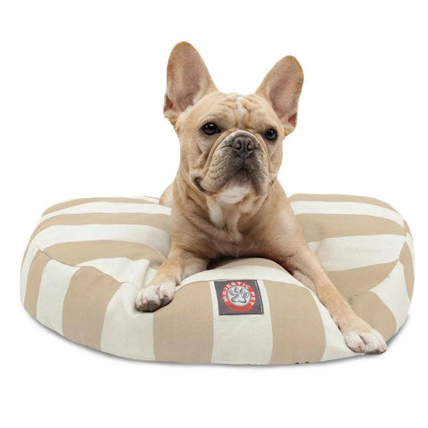 A light brown French Bulldog sits on a Majestic Pet Vertical Stripe Round Dog Bed, featuring beige and white stripes, a logo patch in front, and a removable cover for easy care. The dog looks forward with its ears perked up.