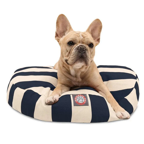 A light brown French Bulldog rests on the Majestic Pet Vertical Stripe Round Dog Bed, featuring navy and white stripes, a removable cover, and a logo patch on the front, gazing forward with its ears perked up.