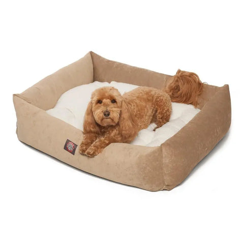 A small, curly-haired brown dog is lying comfortably in the Majestic Pet Villa Luxe Bagel Bed, a rectangular beige bed featuring plush white cushioning.