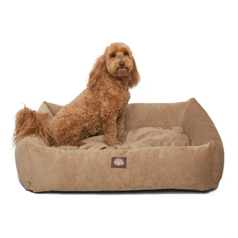 A small, curly-haired brown dog lounges on the Villa Luxe Bagel Bed by Majestic Pet, featuring raised edges and a plush, tan design for ultimate comfort against a white background.