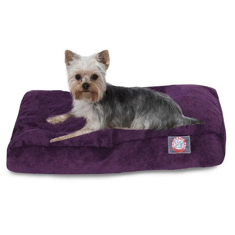 A small dog with gray and tan fur is lying on a plush Villa Rectangle Dog Bed by Majestic Pet, filled with premium polyester fiber fill and featuring a paw print logo patch on the front.