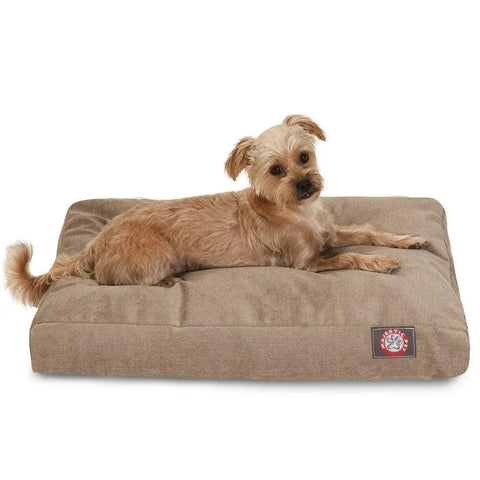 A small, light brown dog with floppy ears rests on the Majestic Pet Villa Memory Foam Dog Bed, which has a washable tan removable cover and a red and white logo patch sewn onto the front corner.