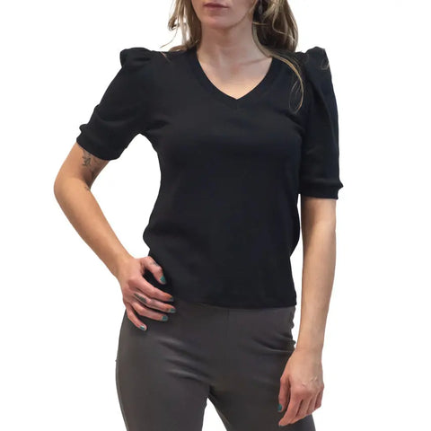 A woman stands confidently with one hand on her hip, wearing the Lamour Top by Spiritex | Sustainable Apparel—a retro V-neck style with puffed shoulders and short sleeves—paired with gray fitted pants against a white background.