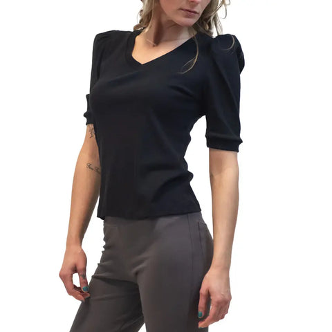 A woman models the Spiritex Lamour Top, a fitted black retro V-neck with puffed sleeves, paired with gray pants. She stands against a white background, turned slightly to the side.