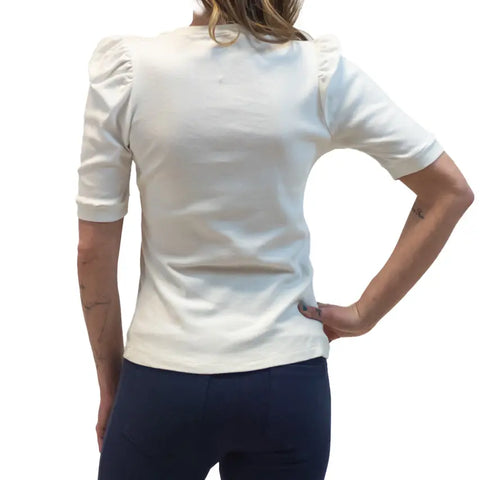A woman stands with her back to the camera, wearing the Lamour Top by Spiritex | Sustainable Apparel—an organic cotton white top with puffed shoulders—paired with dark pants. One hand rests on her hip and her light brown hair is down.
