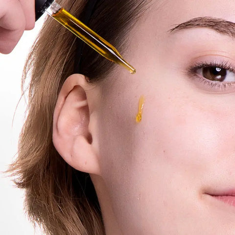 A close-up of a person’s light skin as a dropper applies Open Formula Vitamin C Serum to their cheek. Wearing a black headband and with short hair, they use the serum to target hyperpigmentation and dark spots.