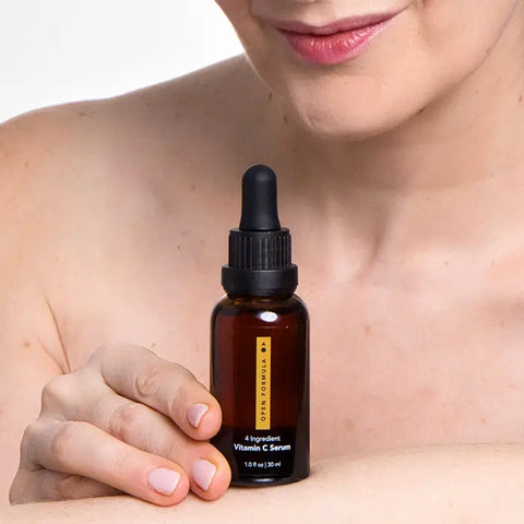 A person with bare shoulders smiles gently, holding an Open Formula Vitamin C Serum bottle close to their chest against a plain, light background—suggesting a skincare routine for dark spots.