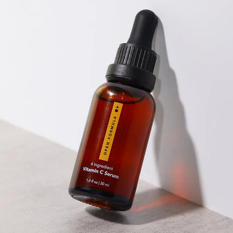 A brown glass dropper bottle of Open Formula Vitamin C Serum with a black cap stands on a white and gray surface. Designed to target dark spots and hyperpigmentation, it casts a subtle shadow.