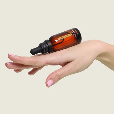 A hand with light skin and manicured nails balances an amber glass dropper bottle of Open Formula Vitamin C Serum—ideal for targeting dark spots—on the back of the fingers, set against a plain beige background.