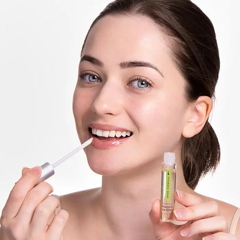 A smiling woman with blue eyes and brown hair pulled back applies Open Formula Vitamin E Lip Gloss to her lips using a wand applicator. She has light skin and is pictured against a plain white background.