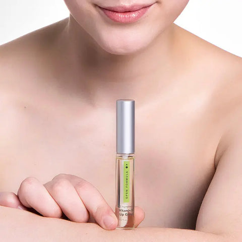A person with bare shoulders holds Open Formula Vitamin E Lip Gloss, a small tube with a silver cap and green label, near their lips against a plain white background.