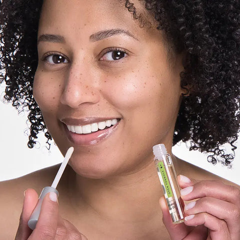 A woman with curly hair smiles as she applies Open Formula Vitamin E Lip Gloss to her lips with a wand, holding the tube in one hand; her complexion looks natural and radiant.