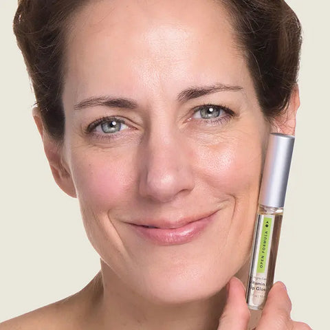 A woman with light skin and blue eyes smiles gently while holding a tube of Open Formula Vitamin E Lip Gloss next to her face.
