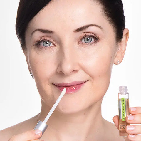 A fair-skinned woman with blue eyes and dark hair smiles as she applies Open Formula Vitamin E Lip Gloss to her lips using a wand, holding the tube in one hand. The background is white.