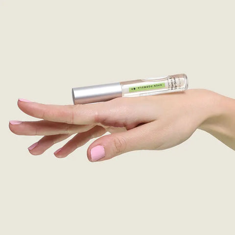 A hand with light pink nails balances a small perfume bottle and an Open Formula Vitamin E Lip Gloss tube against a plain beige background.