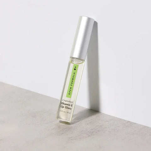 A clear tube of Open Formula Vitamin E Lip Gloss with a silver cap stands upright on a light surface. The label highlights its key ingredients: Vitamin E and Castor Oil.